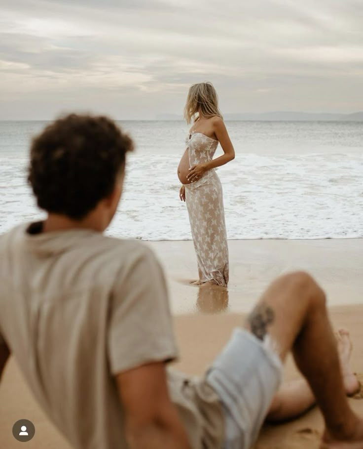 a woman in a white dress standing on the beach next to a man sitting down