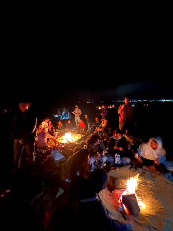 a group of people sitting around a campfire on the beach at night with bright lights