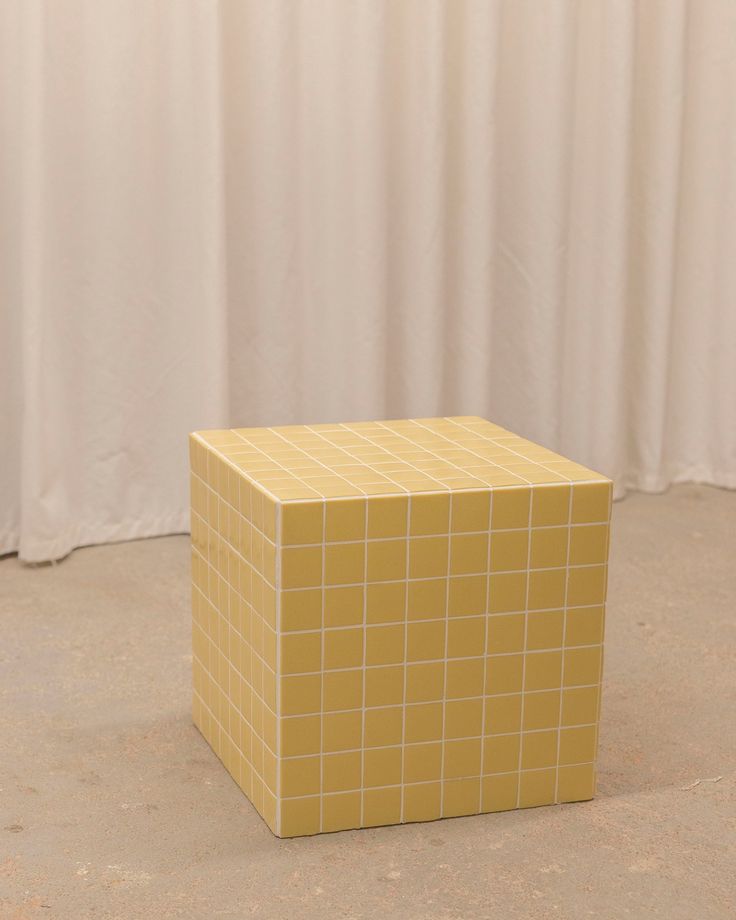 a yellow cube sitting on the ground in front of a white wall with curtains behind it