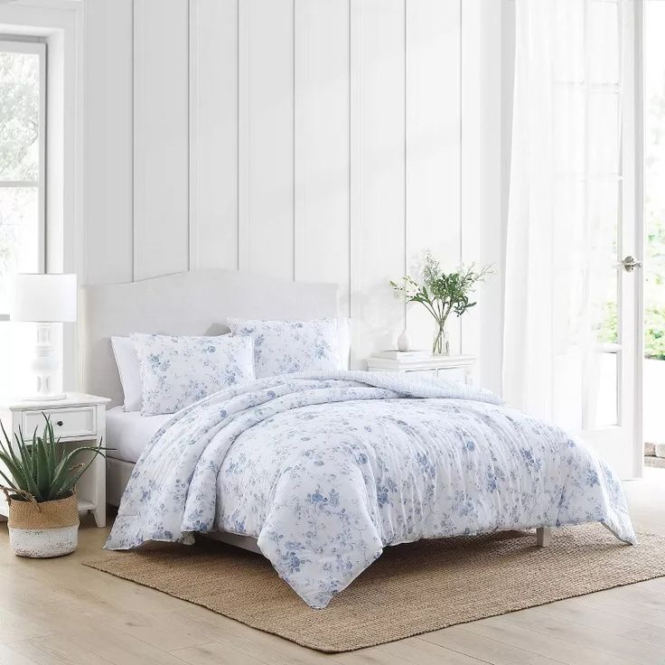 a white bed with blue flowers on the comforter and pillows in front of a window