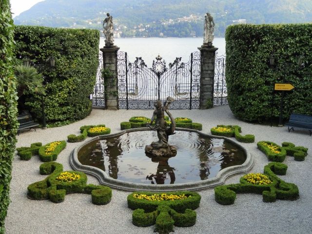 a fountain in the middle of a garden surrounded by hedges and bushes with statues on each side