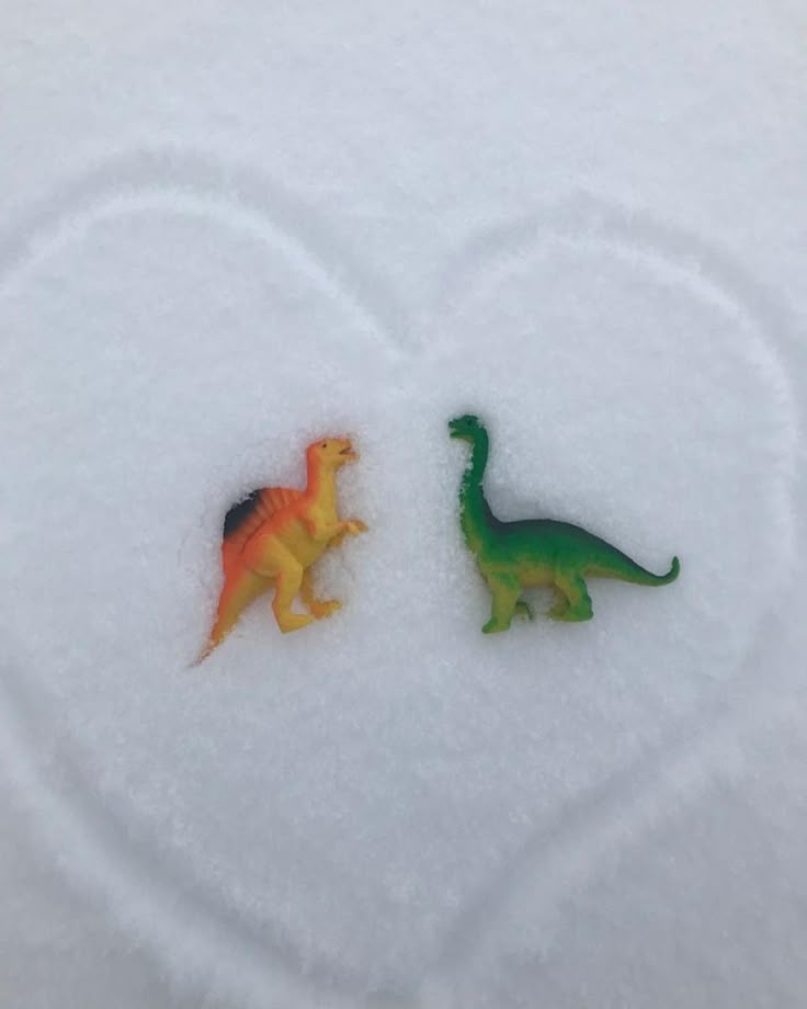 two small plastic dinosaurs in the snow with a heart - shaped hole to make them look like they are facing each other