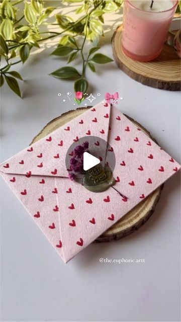 a pink envelope with hearts on it sitting next to a candle and some flowers in the background