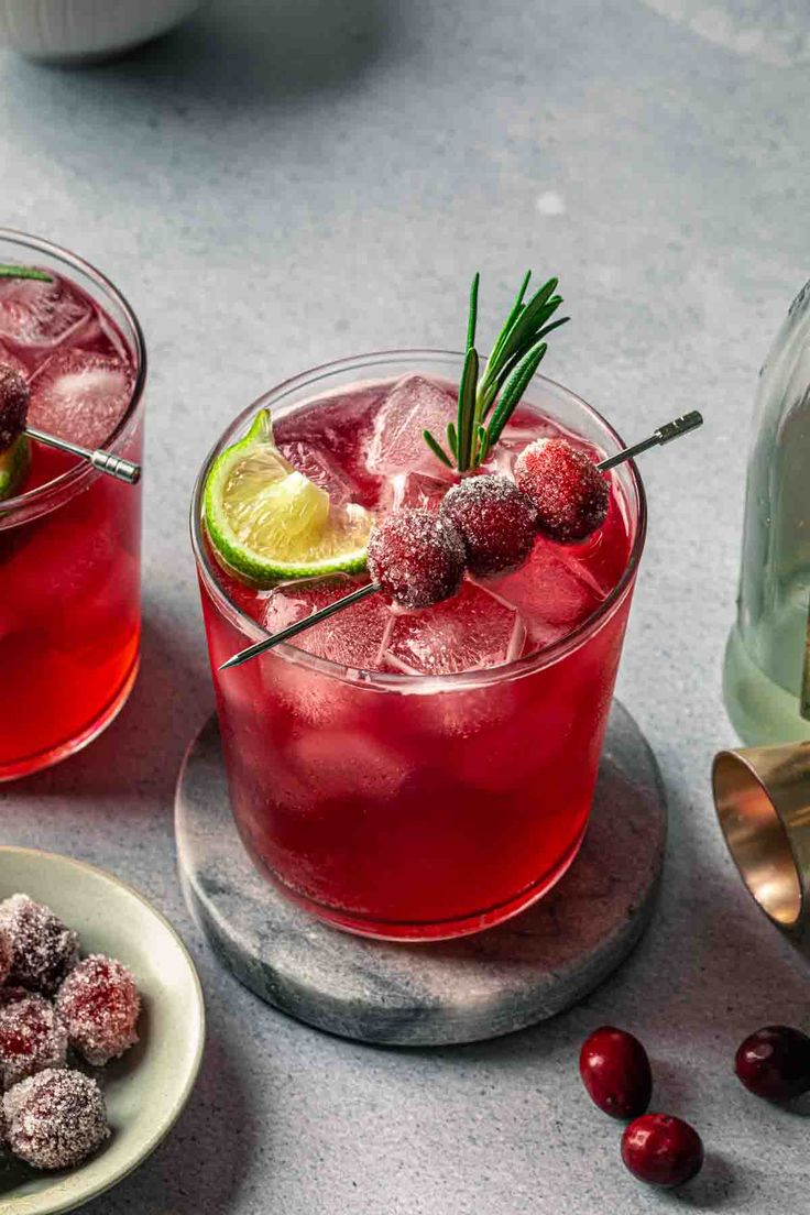 two glasses filled with red liquid and garnished with limes, cranberries and rosemary