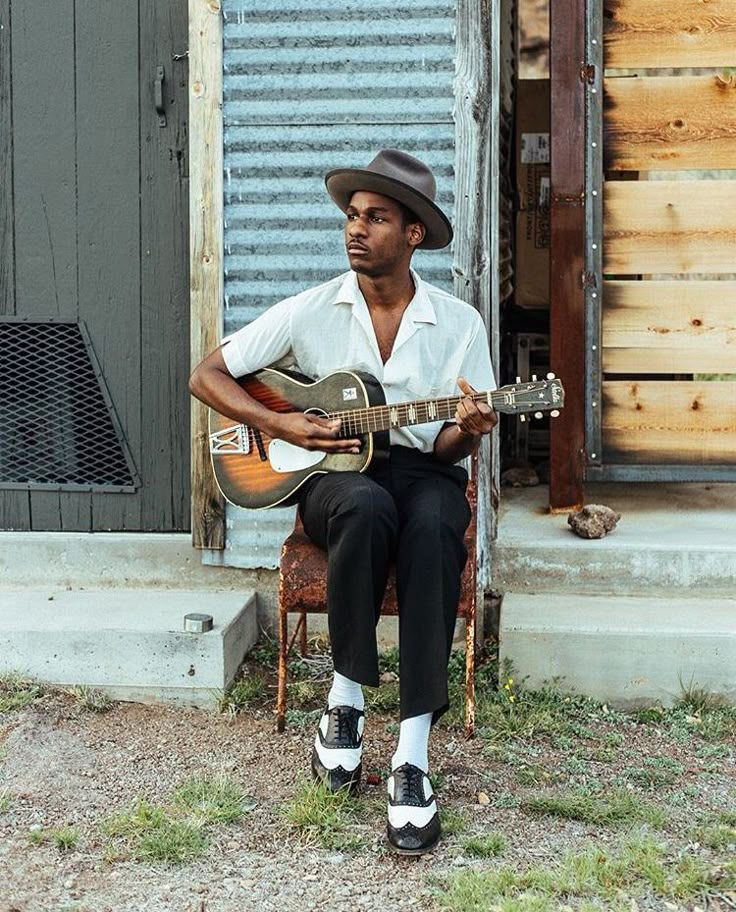 a man sitting on a chair with a guitar in his hand and wearing a hat