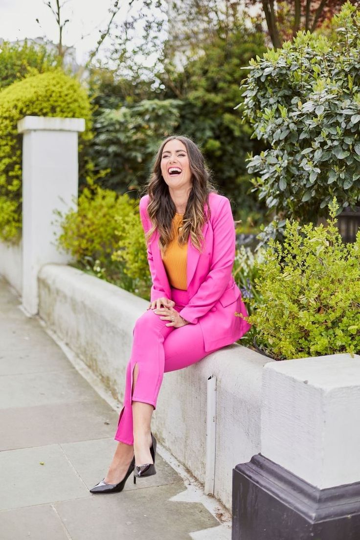 a woman in a pink suit sitting on a cement wall and laughing at the camera