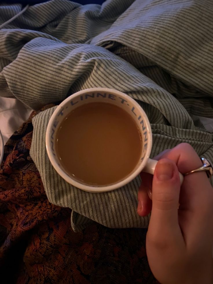 a person holding a cup of coffee on top of a bed next to a blanket