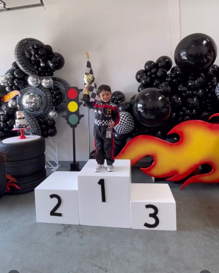 a young boy standing on top of a white pedestal in front of black and red balloons