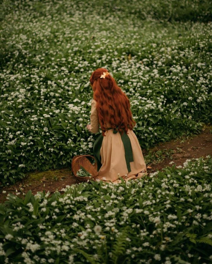 a woman with long red hair sitting on the ground in a field full of white flowers