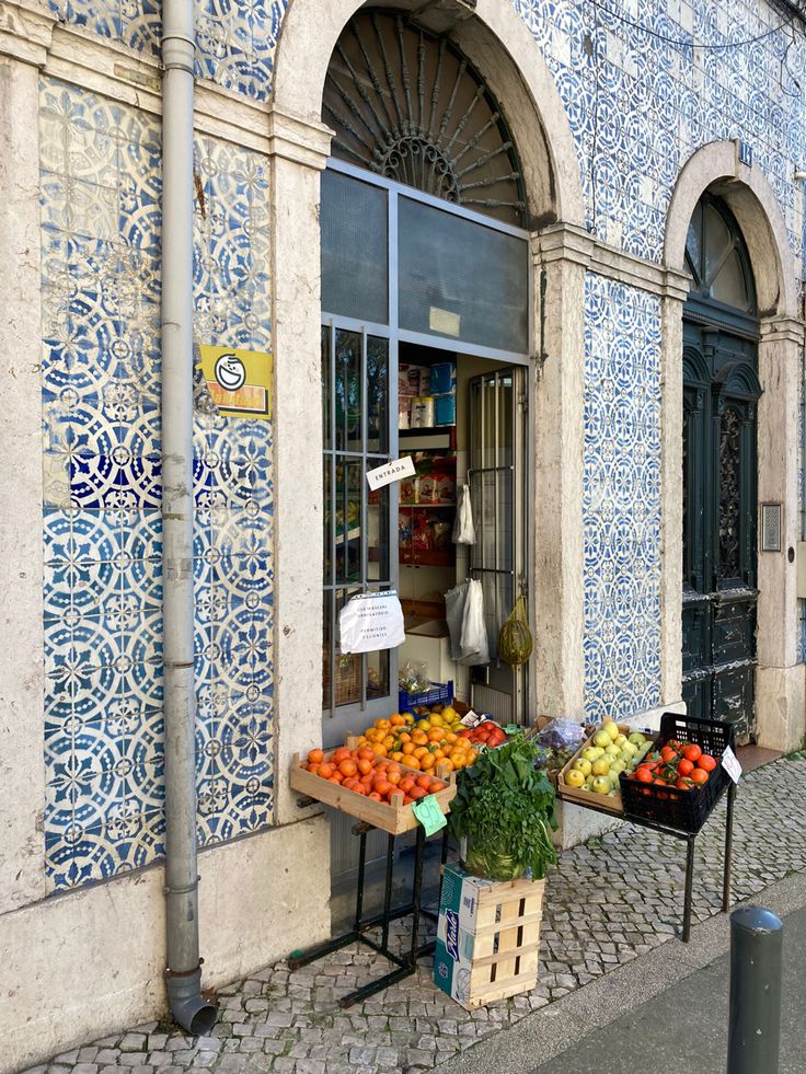 an outdoor fruit and vegetable stand in front of a blue tiled building with arched windows