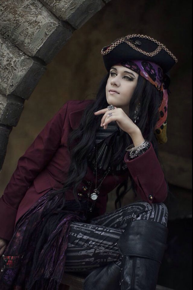 a woman with long black hair wearing a pirate hat and sitting on a stone wall