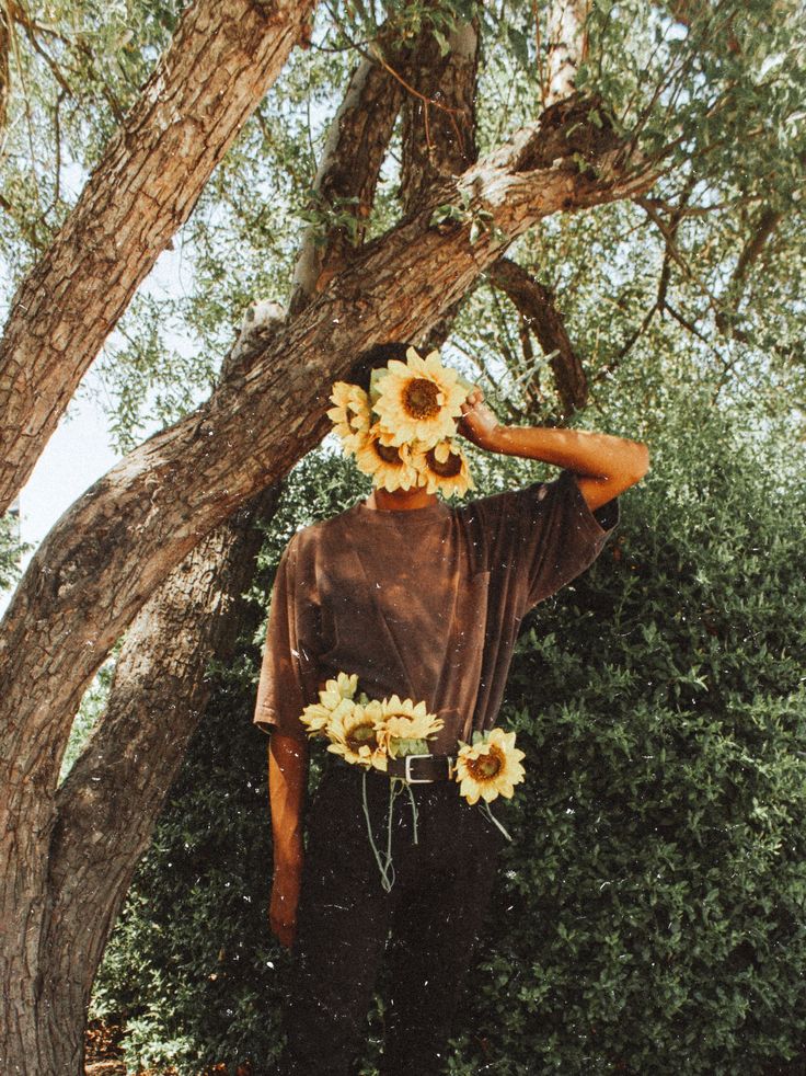 a man with sunflowers on his head standing in front of a large tree