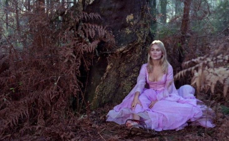 a woman in a pink dress sitting on the ground next to a tree and ferns