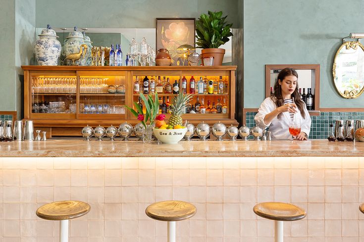 a woman standing behind a bar with three stools in front of it and bottles on the counter