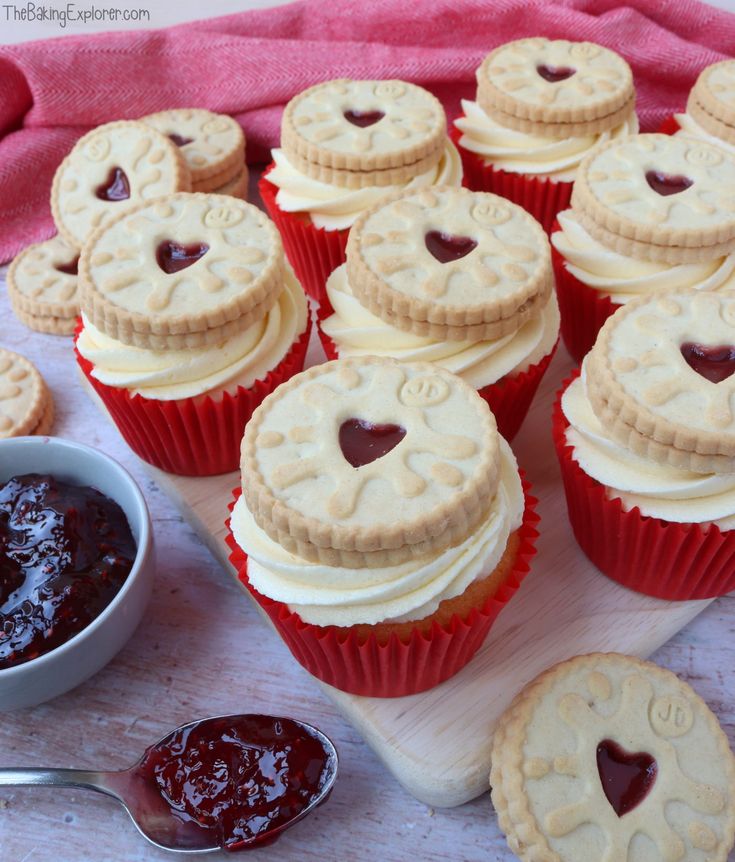 cupcakes with white frosting and heart shaped cookies on a cutting board next to jam