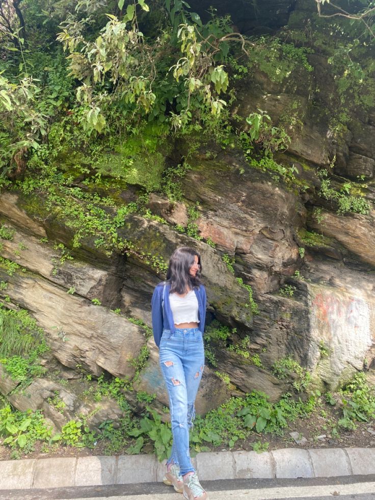 a woman standing on the side of a road next to a rock wall covered in green plants