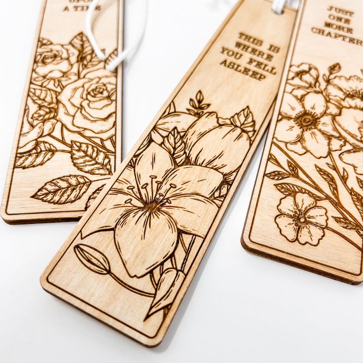 three wooden bookmarks with flowers engraved on the front and back, hanging from strings