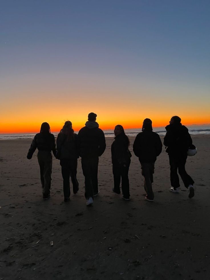 a group of people standing on top of a beach next to the ocean at sunset