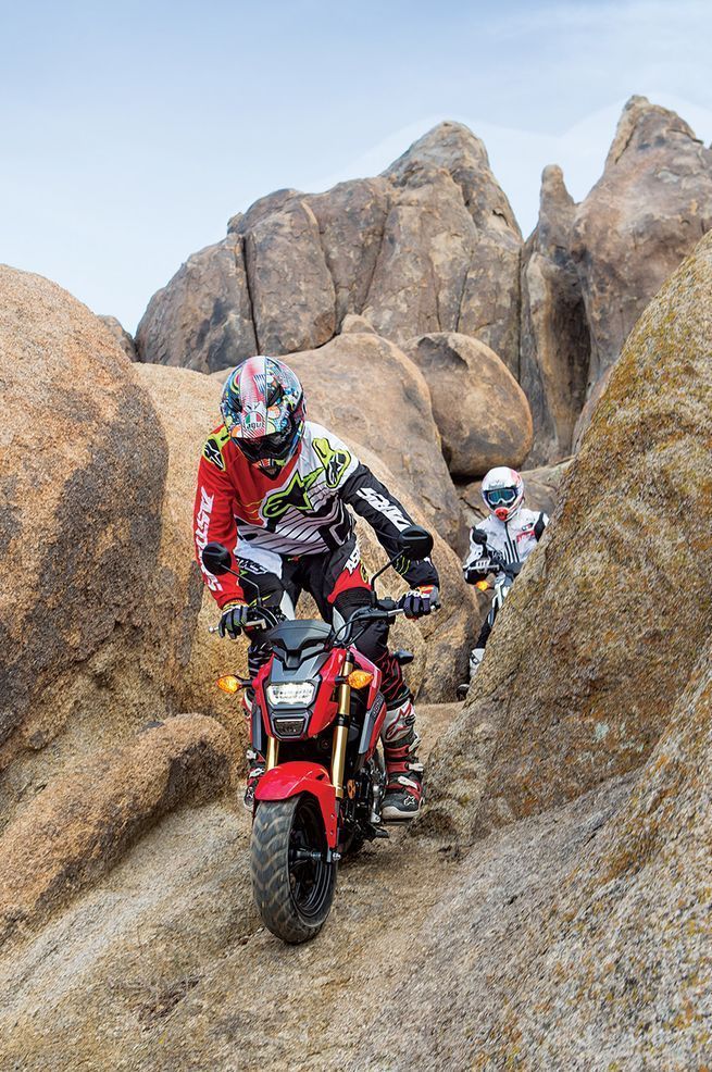 two people on motorbikes going down a rocky trail in the desert with large rocks behind them