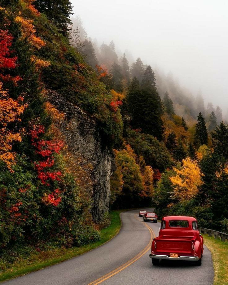 an old red car driving down the road in front of some trees with fall colors