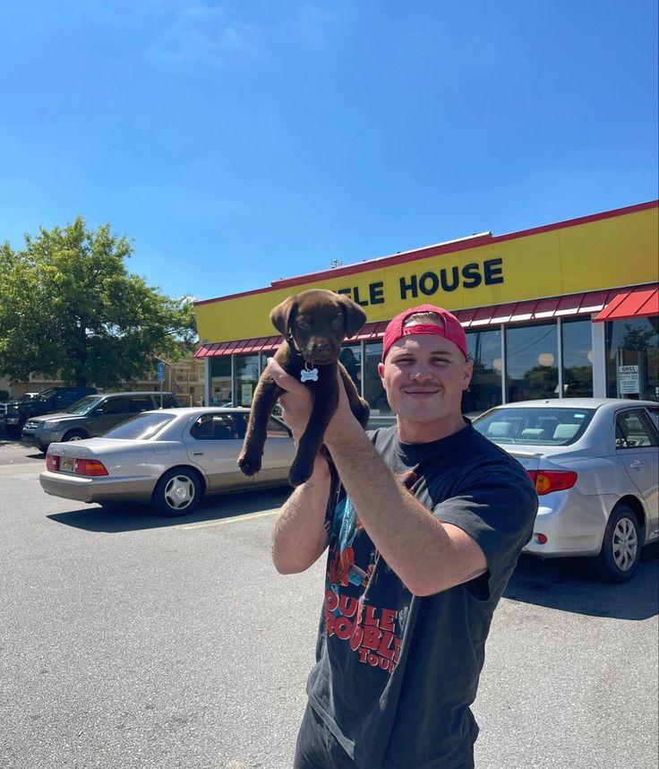 a man holding up a dog in front of a store with cars parked behind him