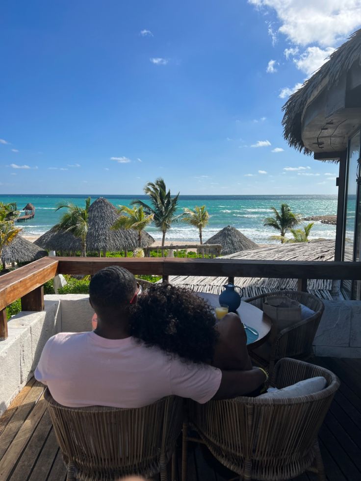 a man and woman sitting on top of a wooden deck next to the ocean with palm trees