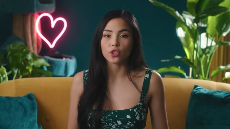 a woman sitting on top of a yellow couch in front of a green wall with a heart shaped neon sign