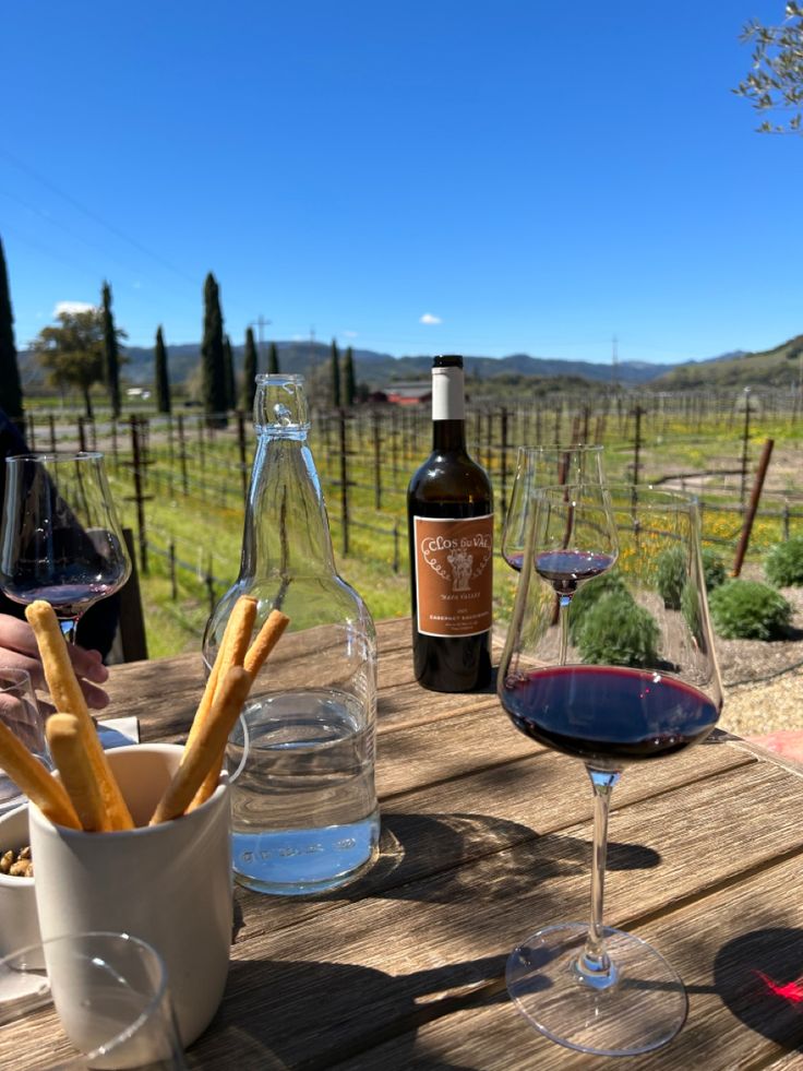 two glasses of wine and some bread sticks on a picnic table with bottles of wine in the background