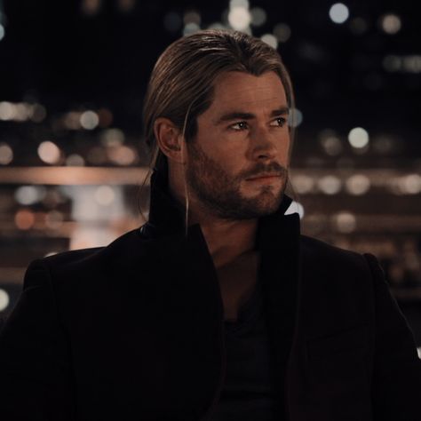 a man with long hair and beard wearing a black jacket looking off into the distance