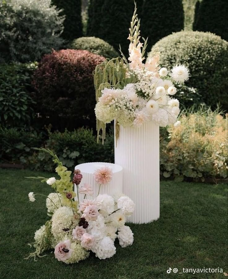 a tall white vase filled with flowers sitting on top of a lush green field next to shrubbery