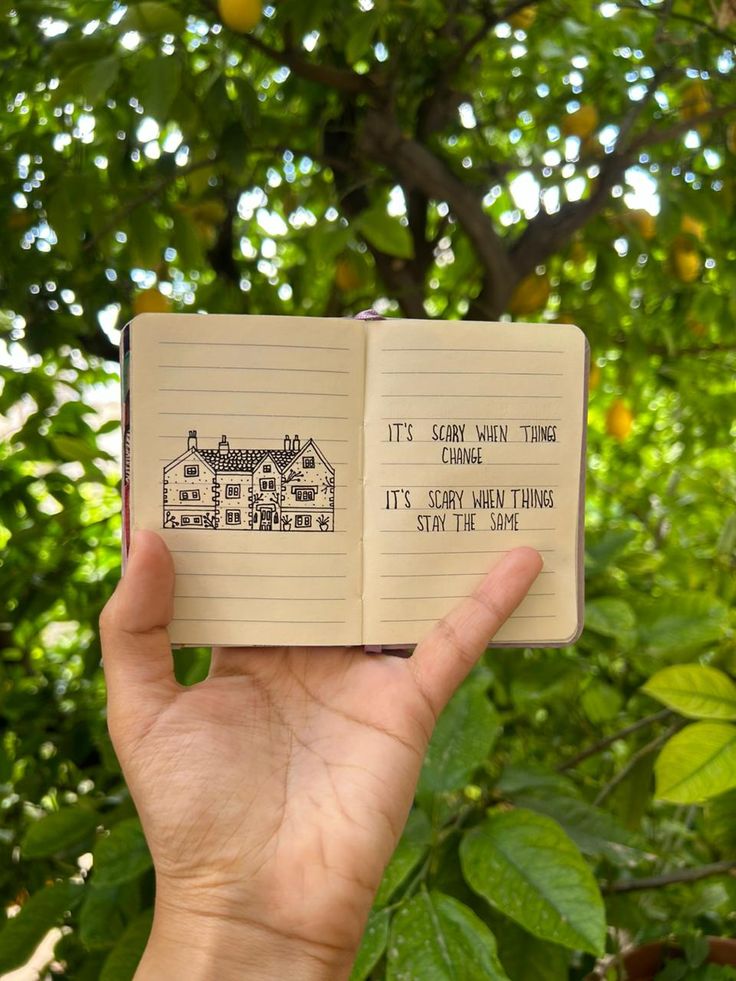 a hand holding an open notebook with writing on it in front of some trees and leaves