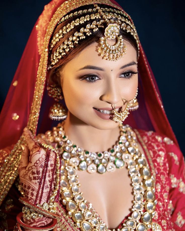 a woman in a red and gold bridal outfit with jewelry on her face, smiling at the camera