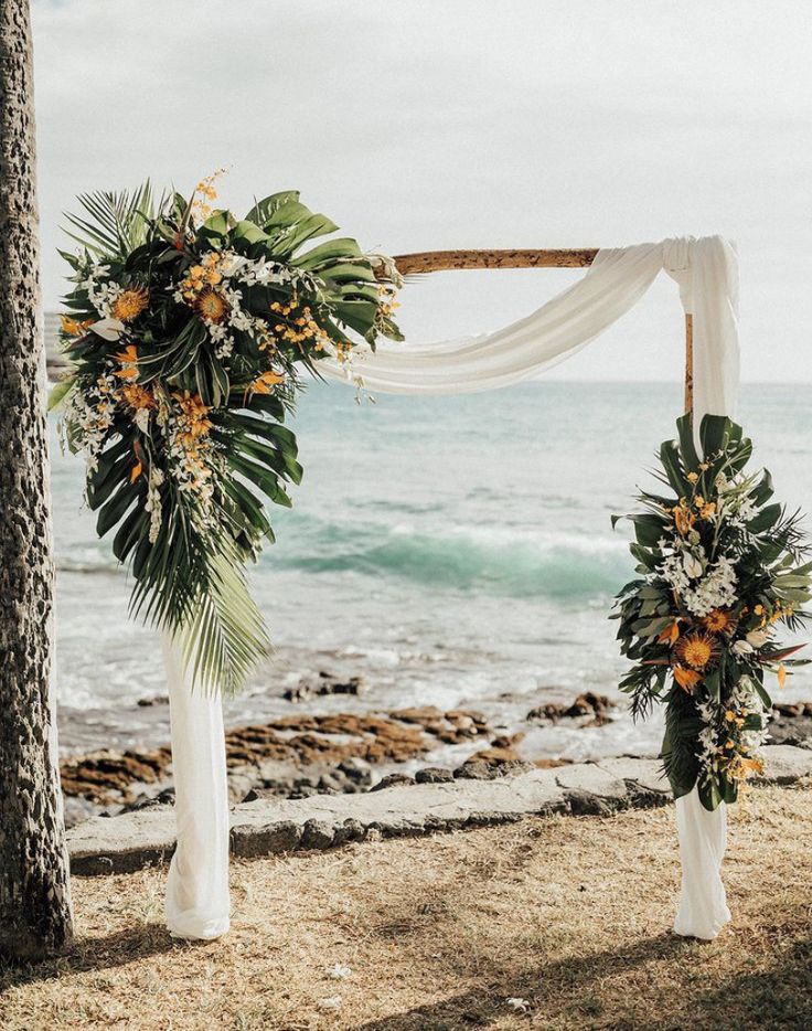 an outdoor wedding setup with white draping and tropical flowers on the arch by the ocean