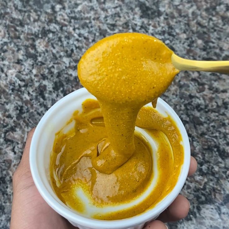 a hand holding a yellow spoon over a white bowl filled with sauce on top of a granite counter