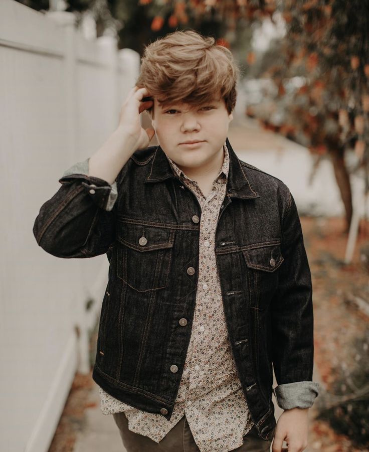 a young man wearing a denim jacket and floral shirt is walking down the sidewalk with his hand on his head