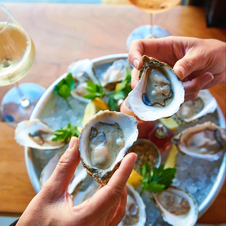 two hands holding oysters on a platter with lemon wedges