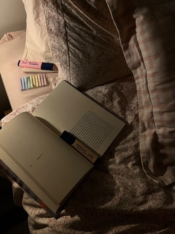 an open book sitting on top of a bed next to some pens and pencils