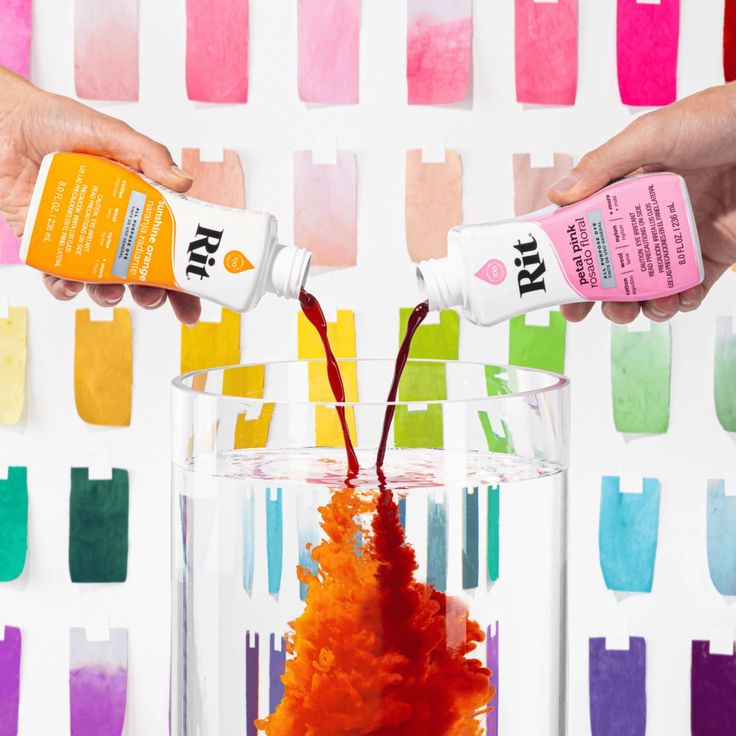 two hands are pouring colored paint into a glass vase filled with orange and red dye