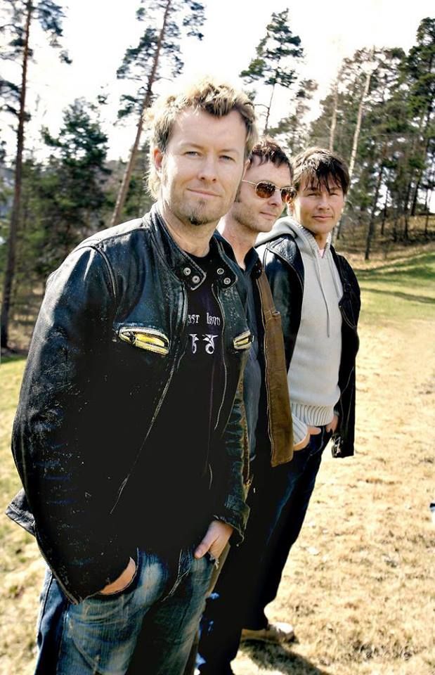 three men standing next to each other in front of trees and grass with one man wearing a leather jacket