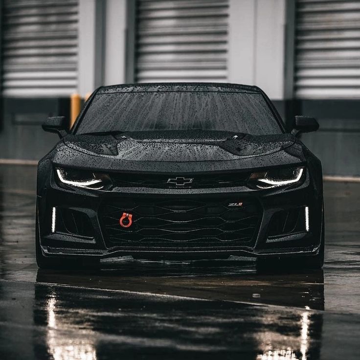 the front end of a black sports car parked on a wet parking lot with garage doors in the background