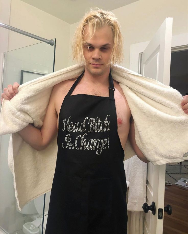 a man with blonde hair wearing an apron and holding a towel over his shoulders in front of a mirror