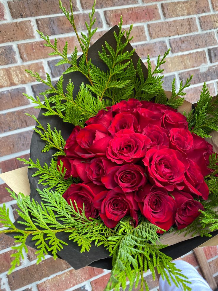 a bouquet of red roses and greenery in front of a brick wall with a person holding it