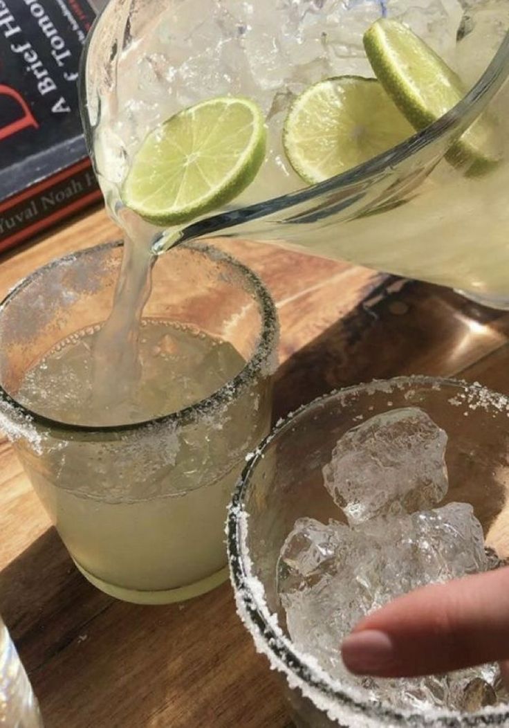 someone is pouring something into two glasses with ice and limes on the table next to them