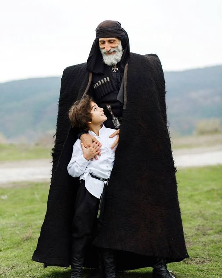 an old man with a child in his lap is posing for the camera while wearing a black cloak