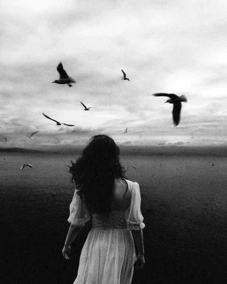 a woman in a white dress walking on the beach with seagulls flying overhead