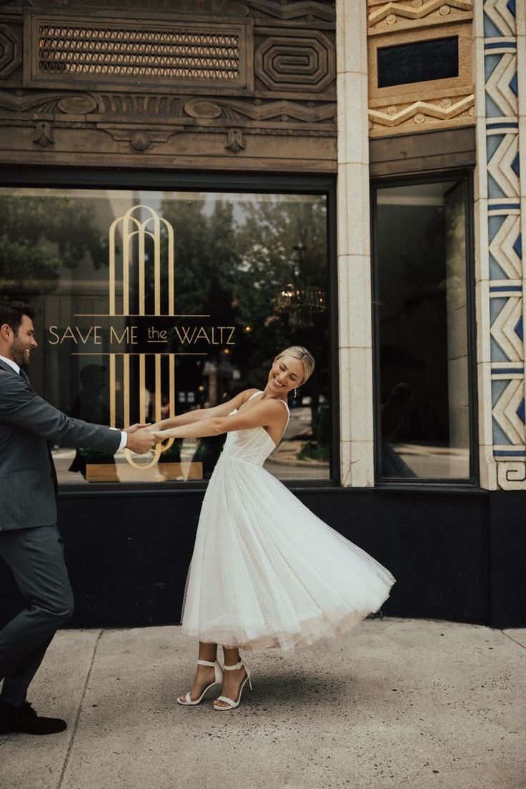 a bride and groom dancing on the sidewalk in front of a storefront with an art deco window behind them