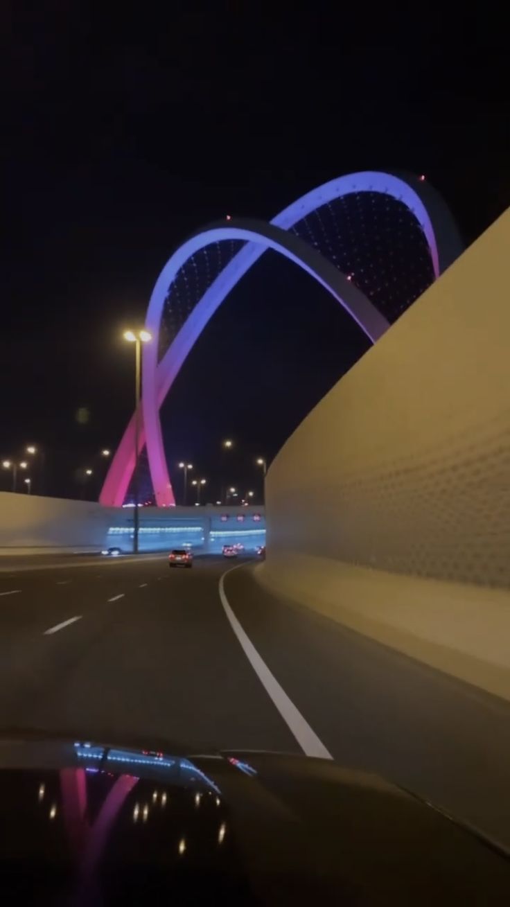 a car driving down a road next to a tall bridge at night with lights on