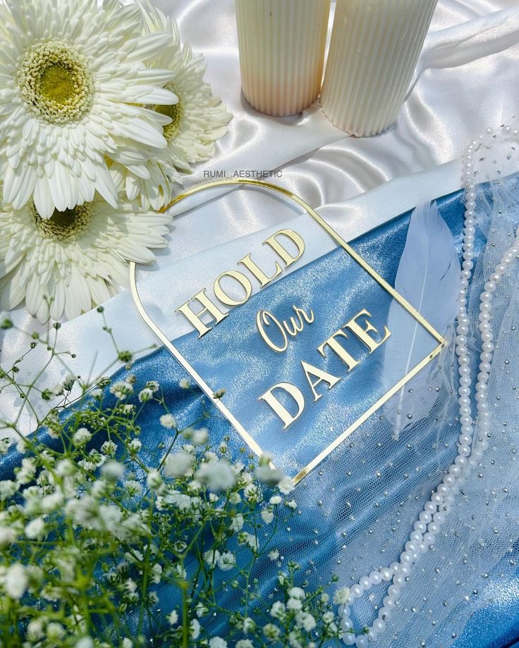 two white flowers sitting next to each other on a blue cloth with the words hold our date