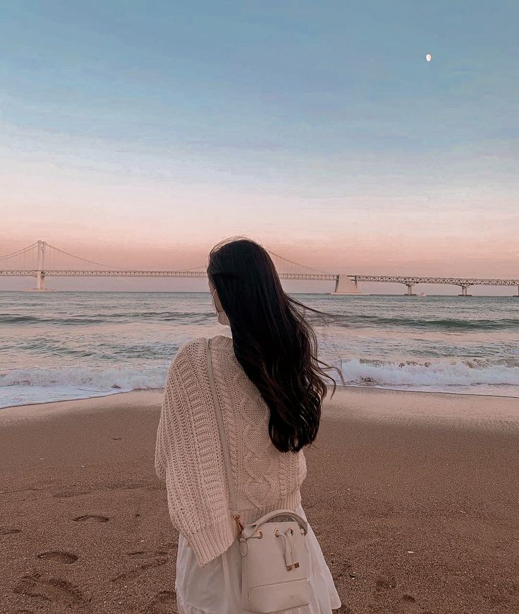 a woman standing on top of a sandy beach next to the ocean with a bridge in the background