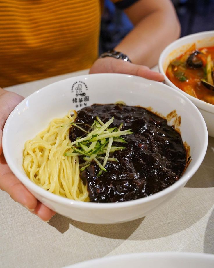 a person holding a bowl of food with noodles and sauce on the table in front of them